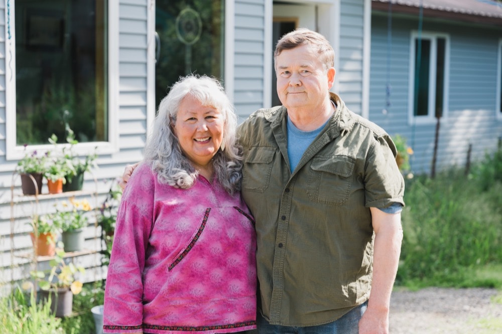 An elder couple stands smiling together outside their home, with the woman wearing a pink tie-dye shirt and the man in a green cargo shirt, set against a backdrop of their cozy house and flourishing garden.