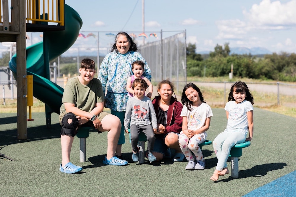 A group of six people, a mix of children and adults, smiling on a sunny day at a playground with slides in the background and a clear blue sky overhead. The group appears joyful and relaxed in a casual outdoor setting.