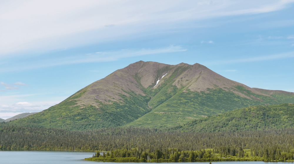 A serene landscape showcasing a towering mountain with lush green slopes under a clear blue sky, flanked by dense forests and a calm lake in the foreground.